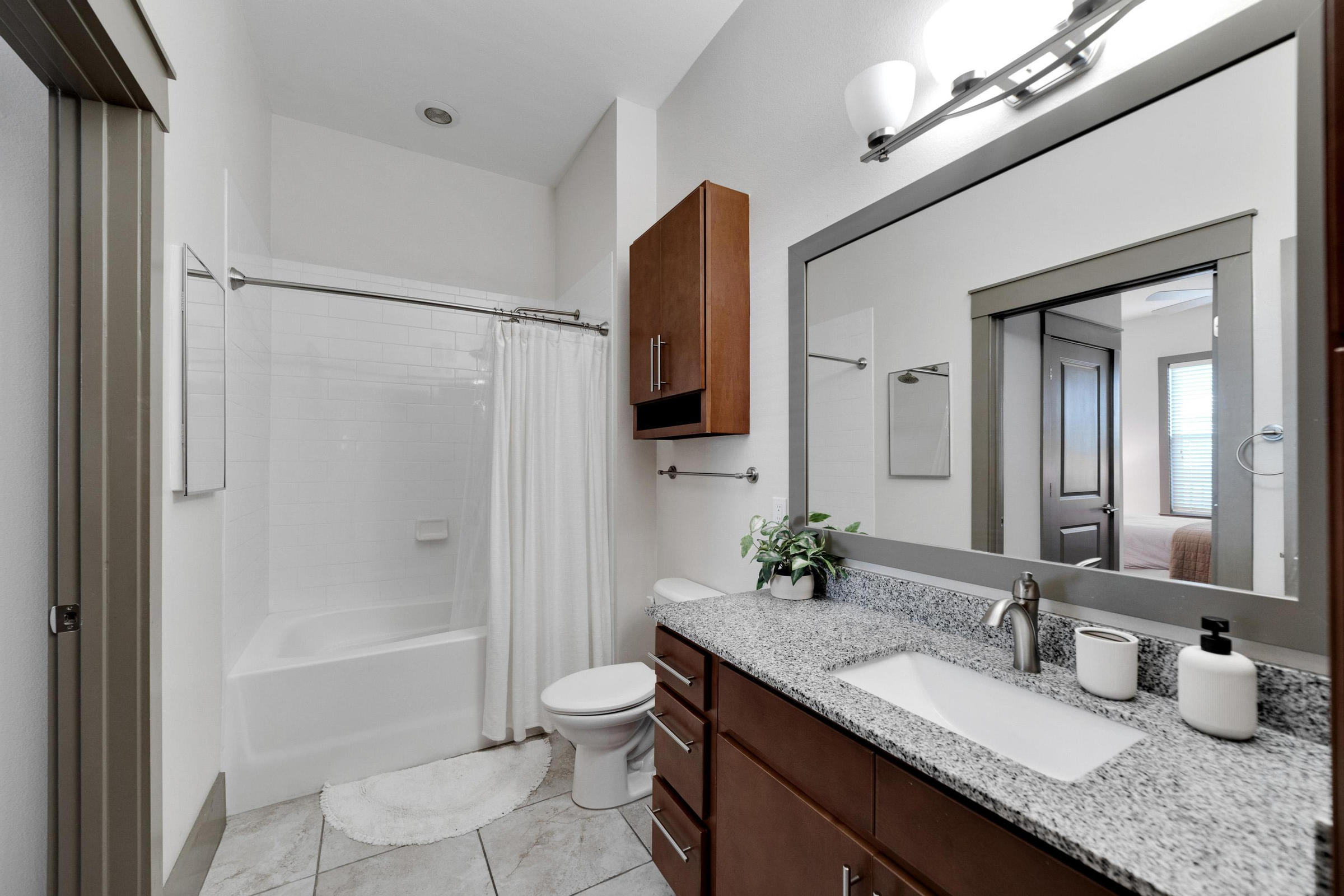 Modern bathroom with granite countertop, wooden cabinets, white shower curtain, and a large mirror above the sink.