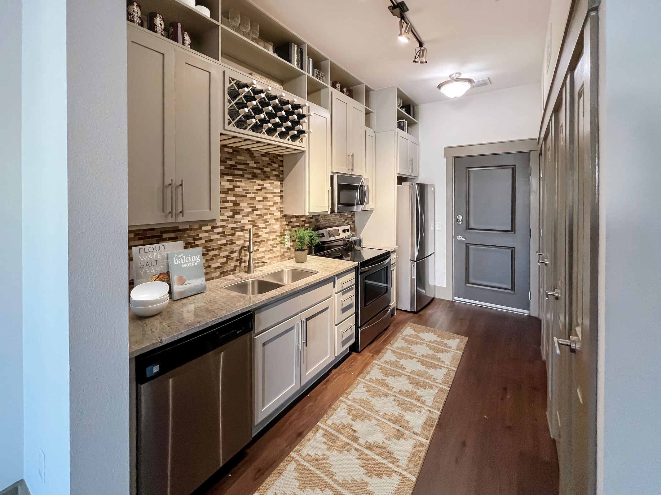 Modern galley kitchen with beige cabinets, stainless steel appliances, a patterned rug, and mosaic tile backsplash.