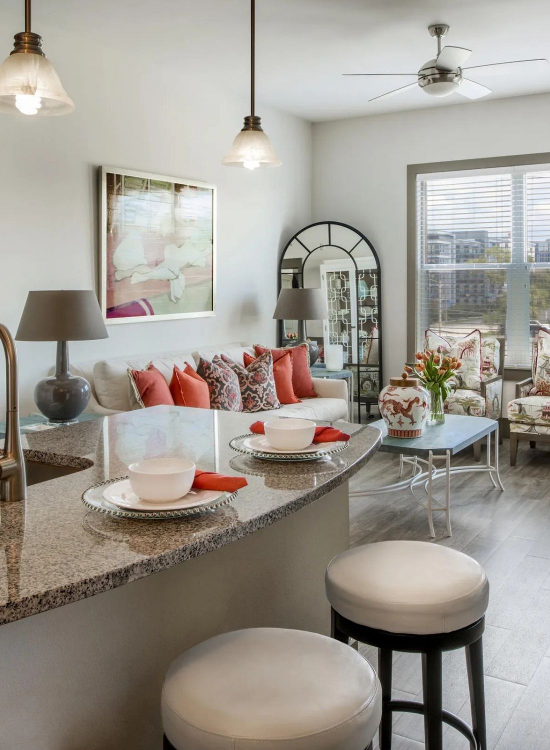Modern living room with large windows, colorful pillows, floral chairs, and a kitchen island with stools in front.