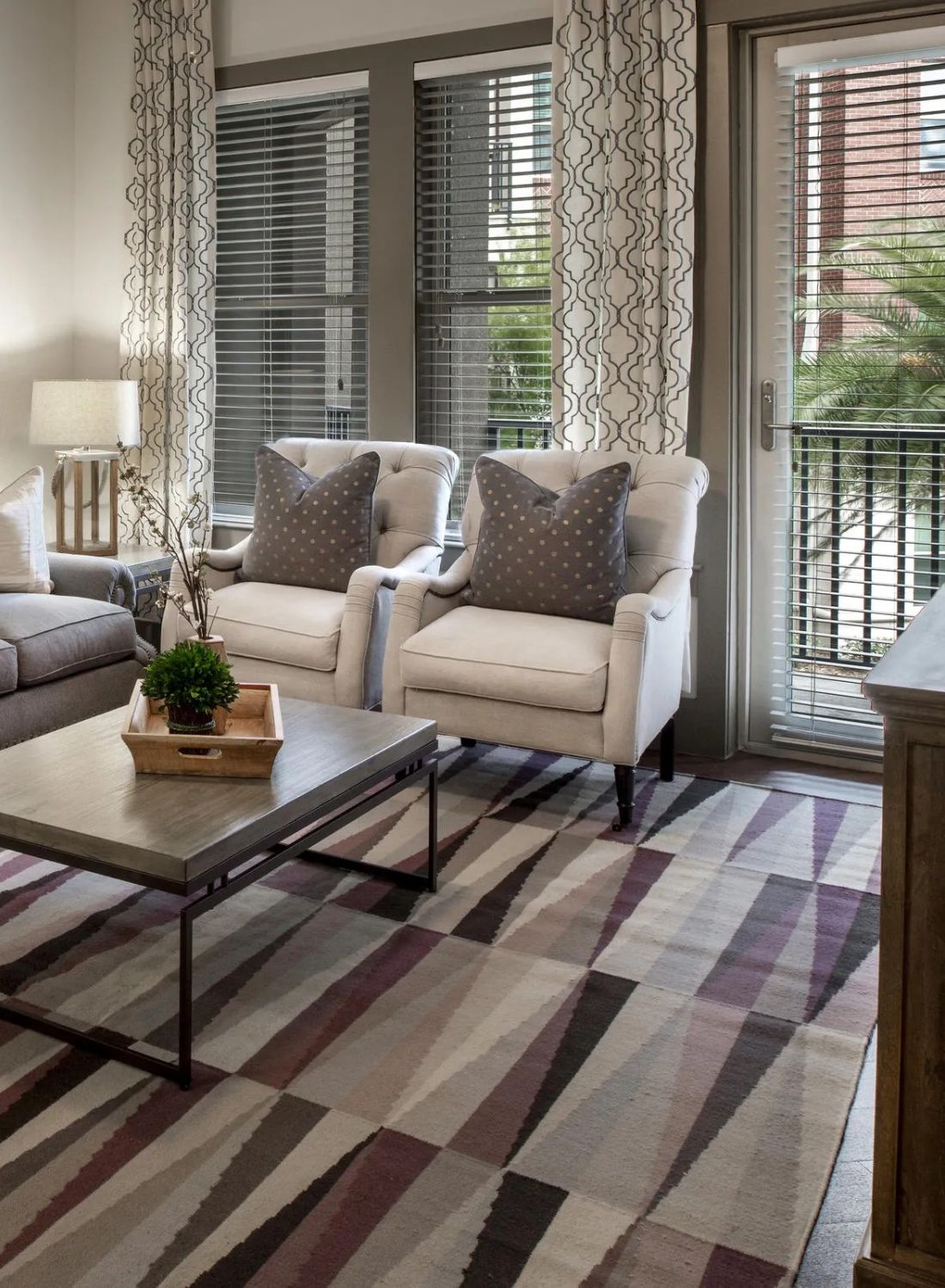Modern living room with a gray sofa, two chairs, patterned rug, coffee table, and large windows with curtains.