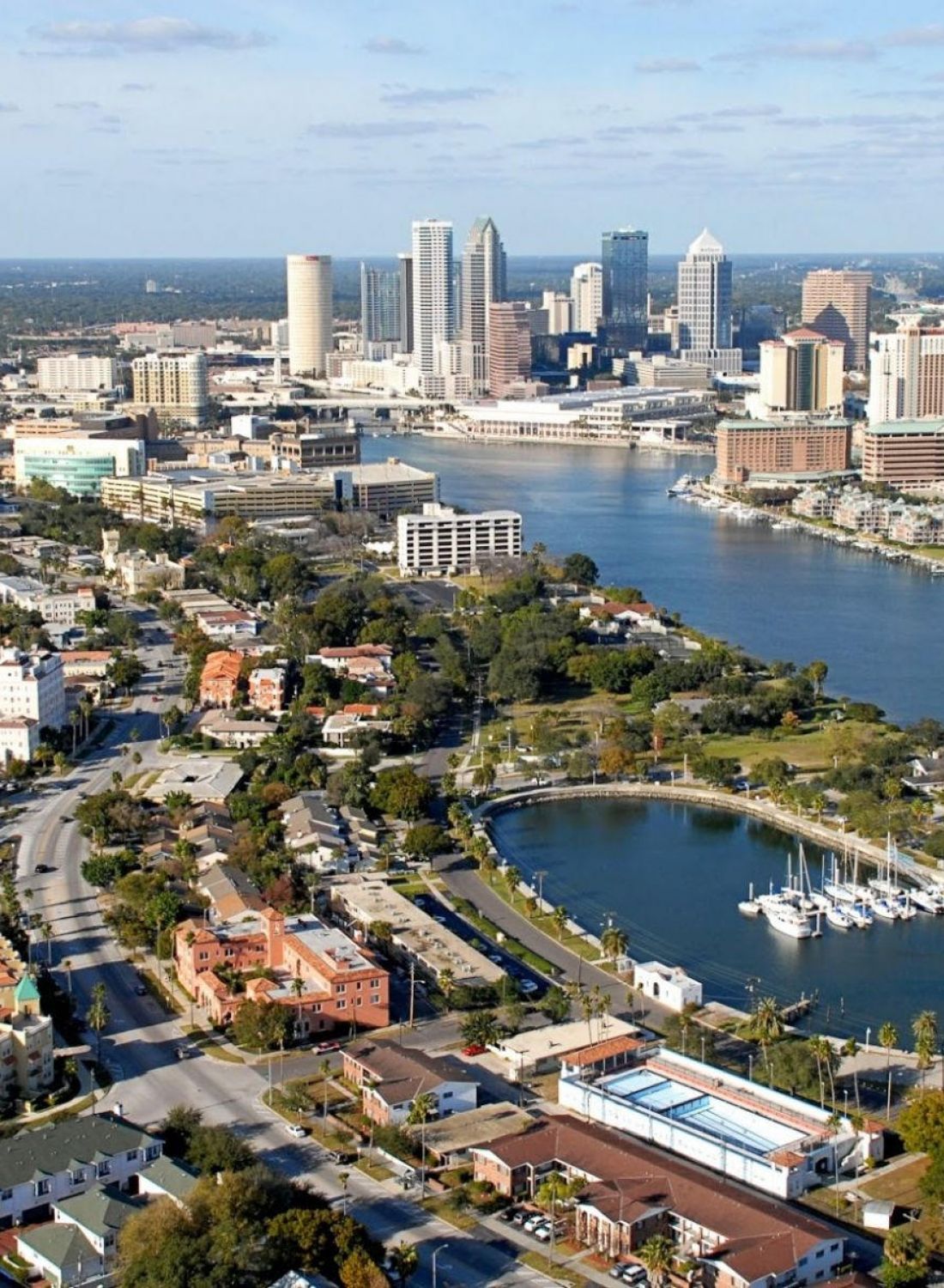 Aerial view of a city skyline with skyscrapers, waterfront, and residential areas under a partly cloudy sky.