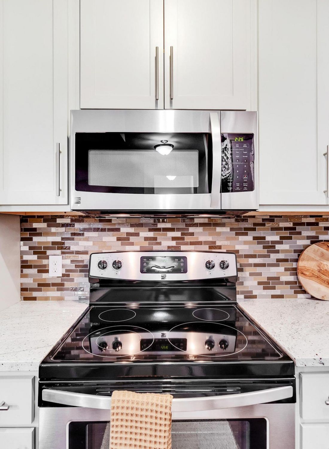 Modern kitchen with white cabinets, stainless steel stove and microwave, mosaic tile backsplash, and a cutting board.