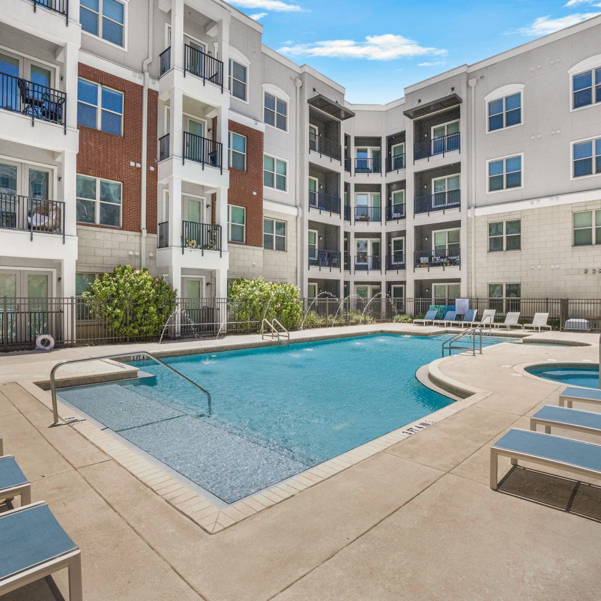 Modern apartment complex with a swimming pool, lounge chairs, and a hot tub on a sunny day.