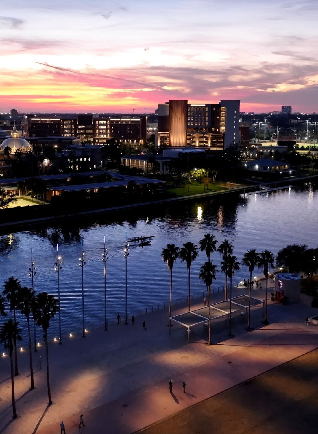 A cityscape at sunset with buildings, a river, palm trees, and people walking along a lit waterfront path.