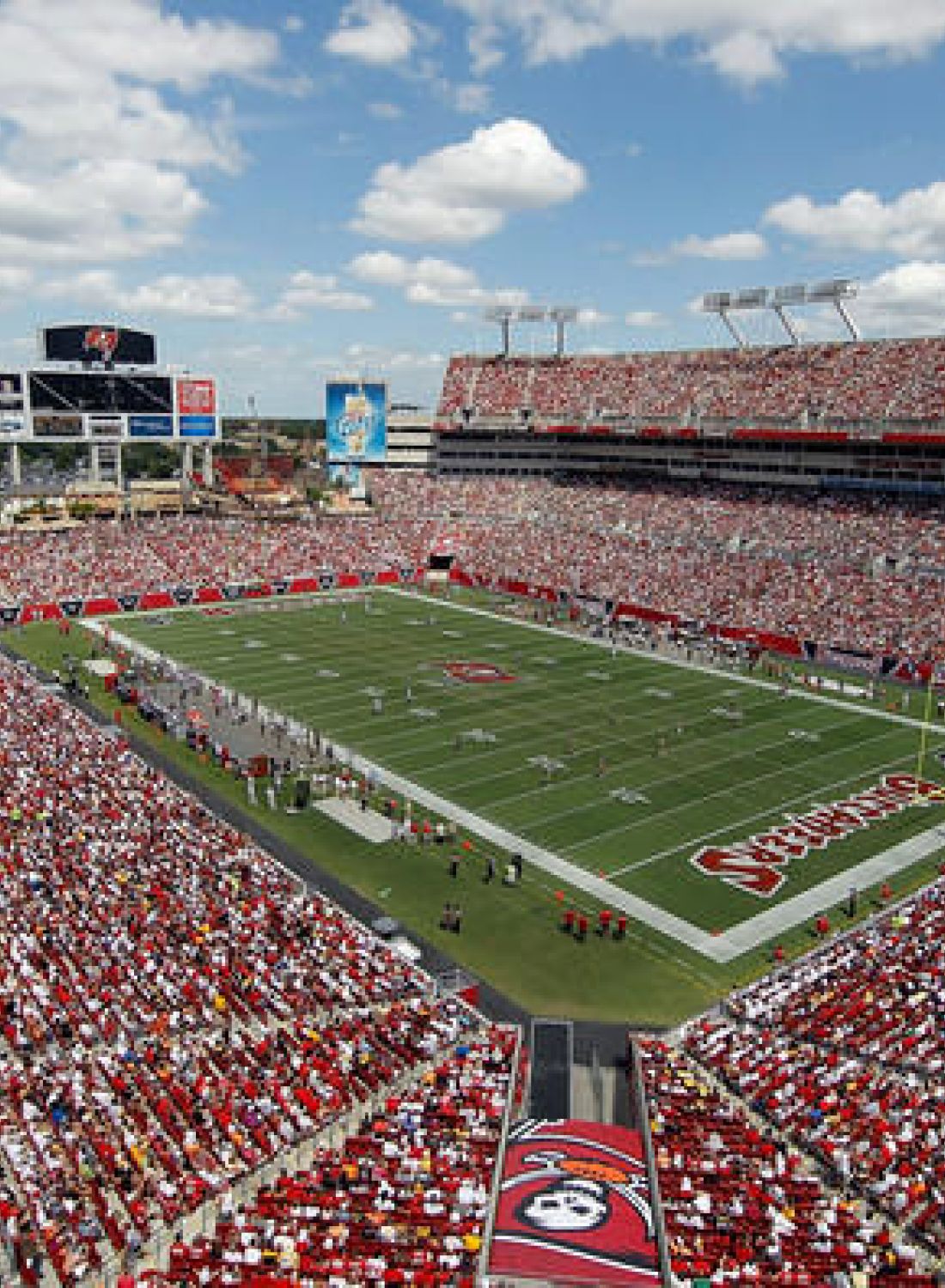 A full football stadium with a game in progress, crowded stands, and partly cloudy skies above.
